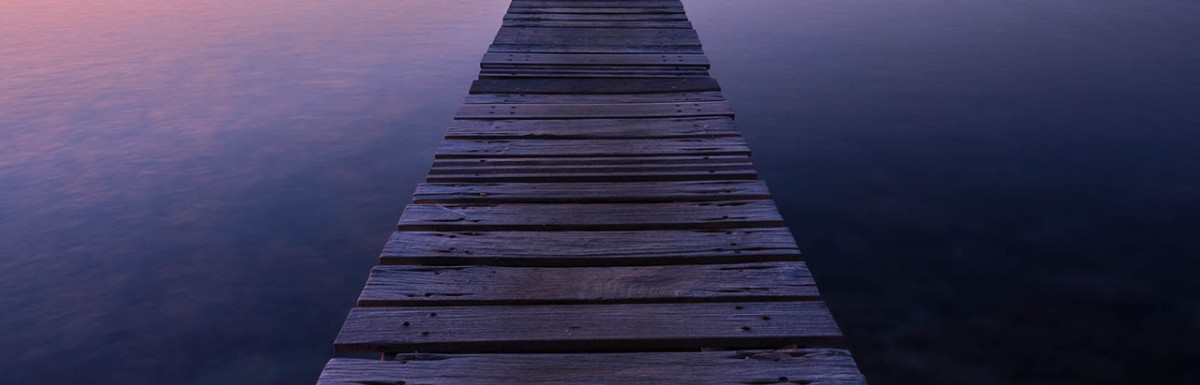 Ein alter Holzsteg, der in einen ruhigen See hineinragt und in der Ferne einen farbenfrohen Sonnenuntergang einfängt.